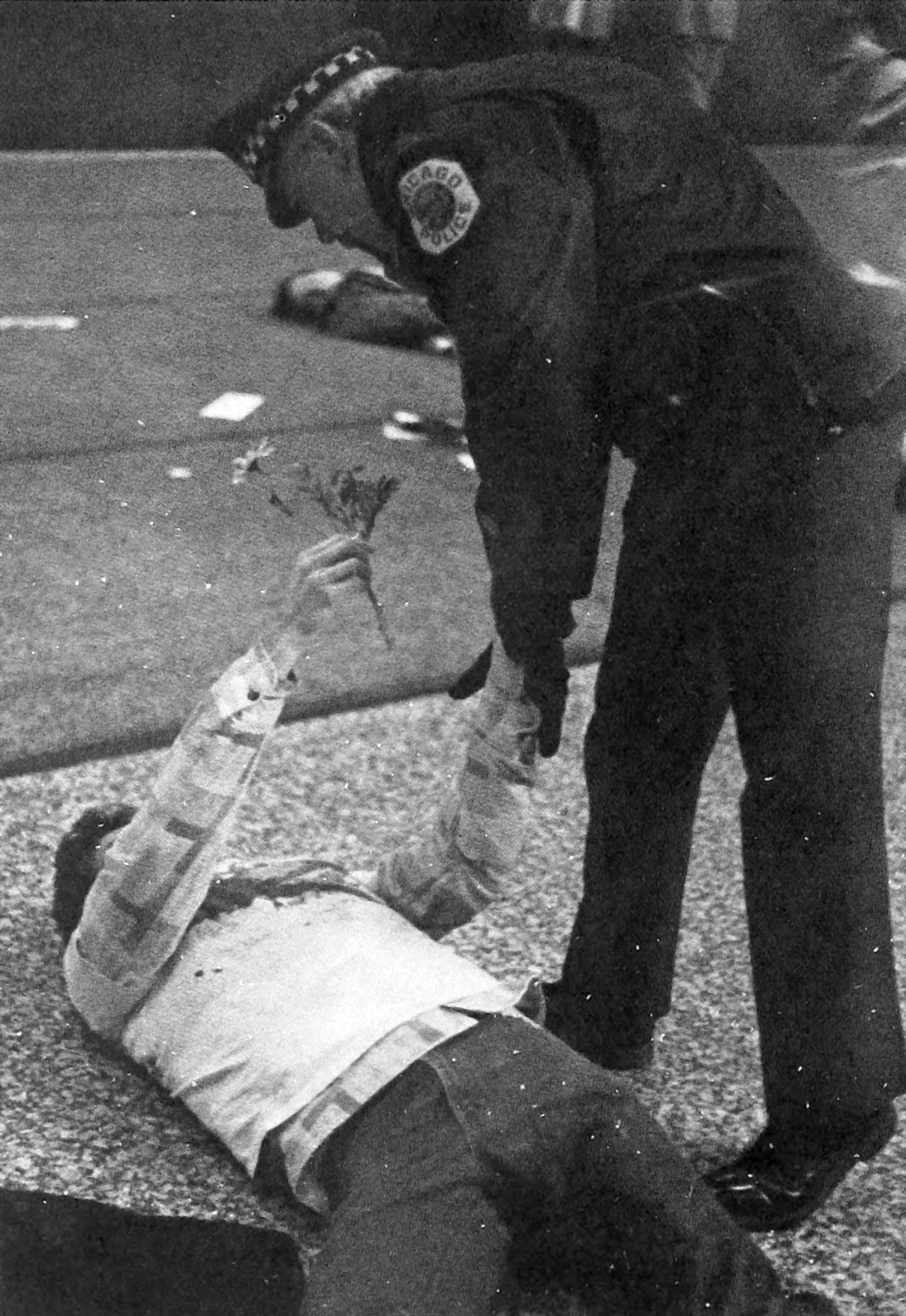 a standing Chicago Police officer grabs one arm of a man lying on his back while with the man’s other hand he offers the officer a flower.