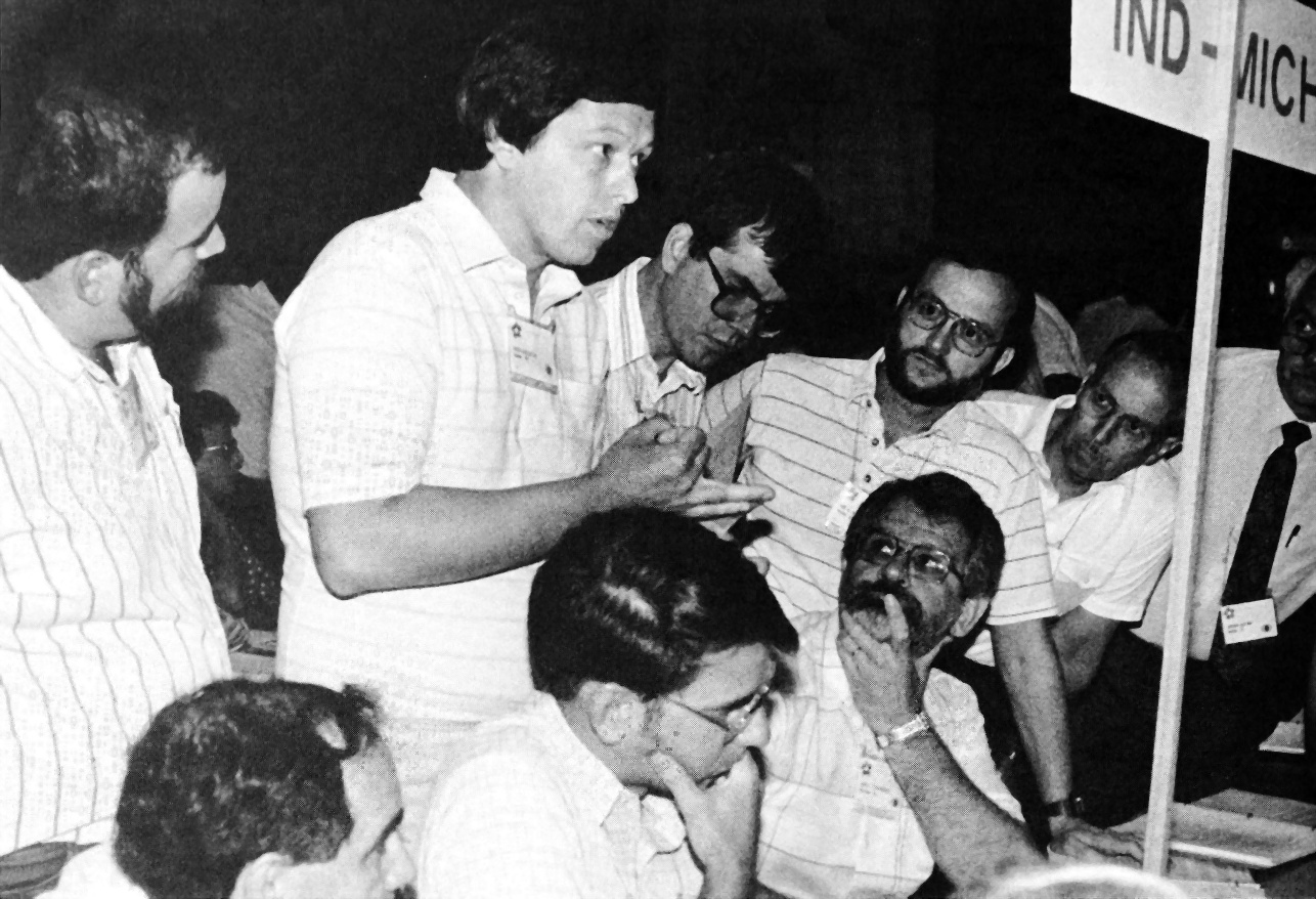 In a group of men, some seated, some standing, Dave Miller gesticulates while he speaks. A sign nearby indicates the table of the Indiana/Michigan delegates.