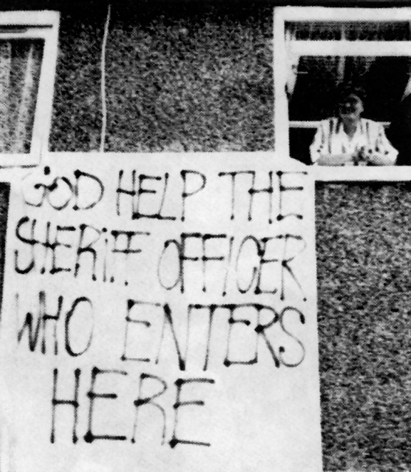 A woman looks out an upstairs window of a house viewed from outside. A hand-painted banner hanging by the window reads “God help the sheriff officer who enters here.”