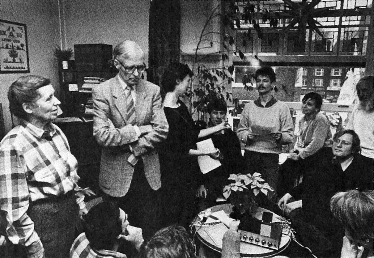 H.B. Kossen, arms crossed, standing, amid a circle of people indoors