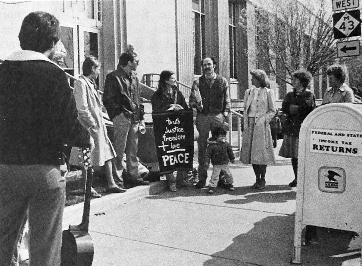 Several people are standing on a sidewalk near mailboxes. One holds a sign reading “truth + justice + freedom + love = peace”.