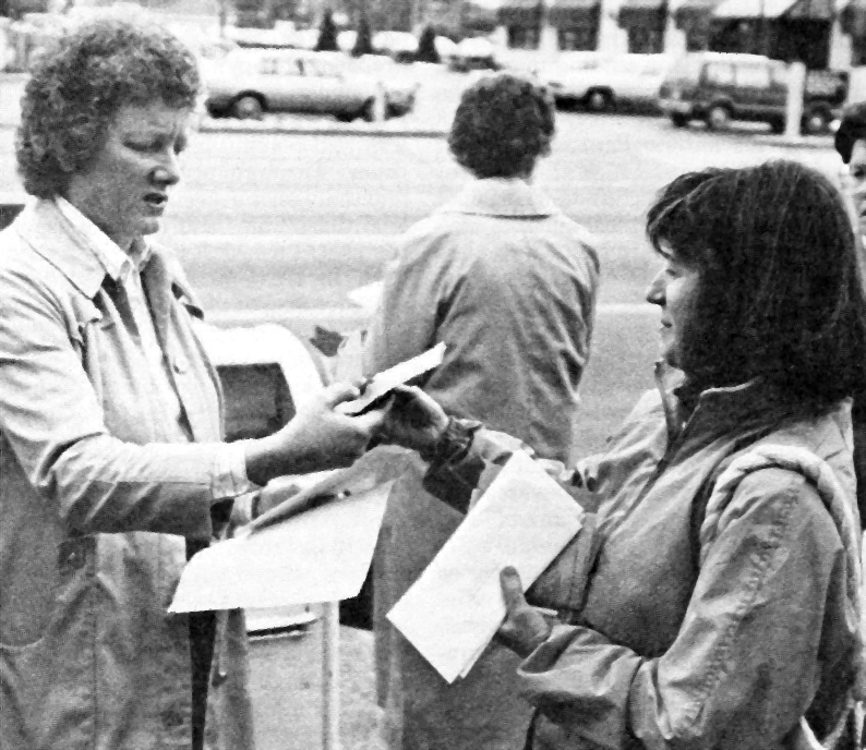 A woman holding envelopes hands a piece of paper to another woman who is holding several sheets of paper. Both women are outside on a sidewalk, wearing jackets. A mailbox is visible behind them.