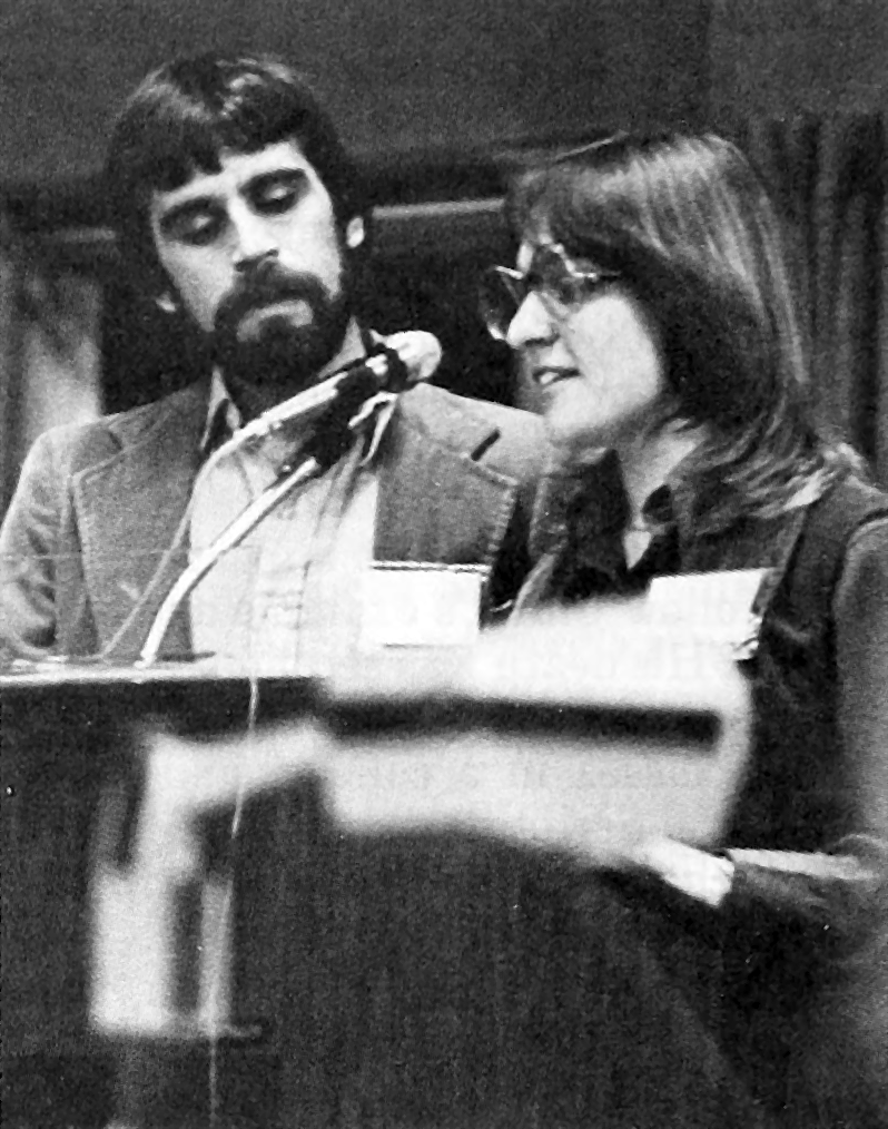 Sandy Drescher Lehman stands at a lectern speaking into a microphone and John Lehman stands nearby.