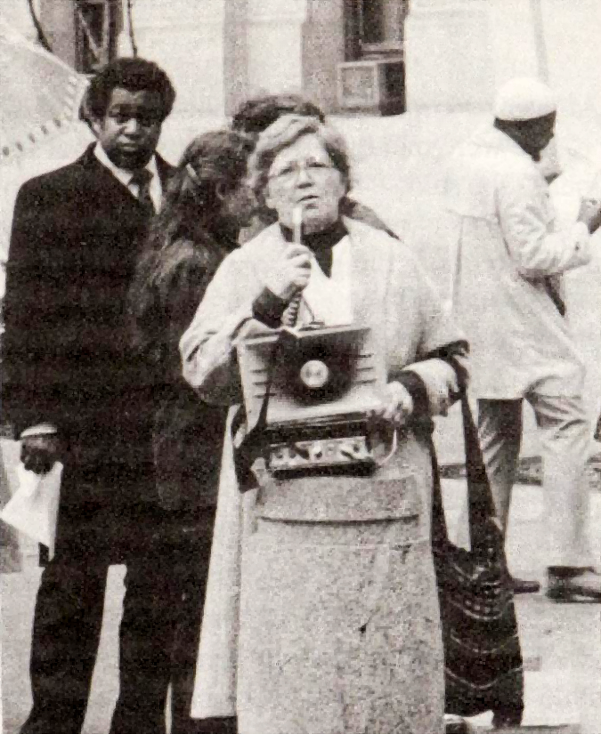Lillian Willoughby speaking into a microphone behind a portable speaker that sits on a concrete bollard outdoors while others stand behind her
