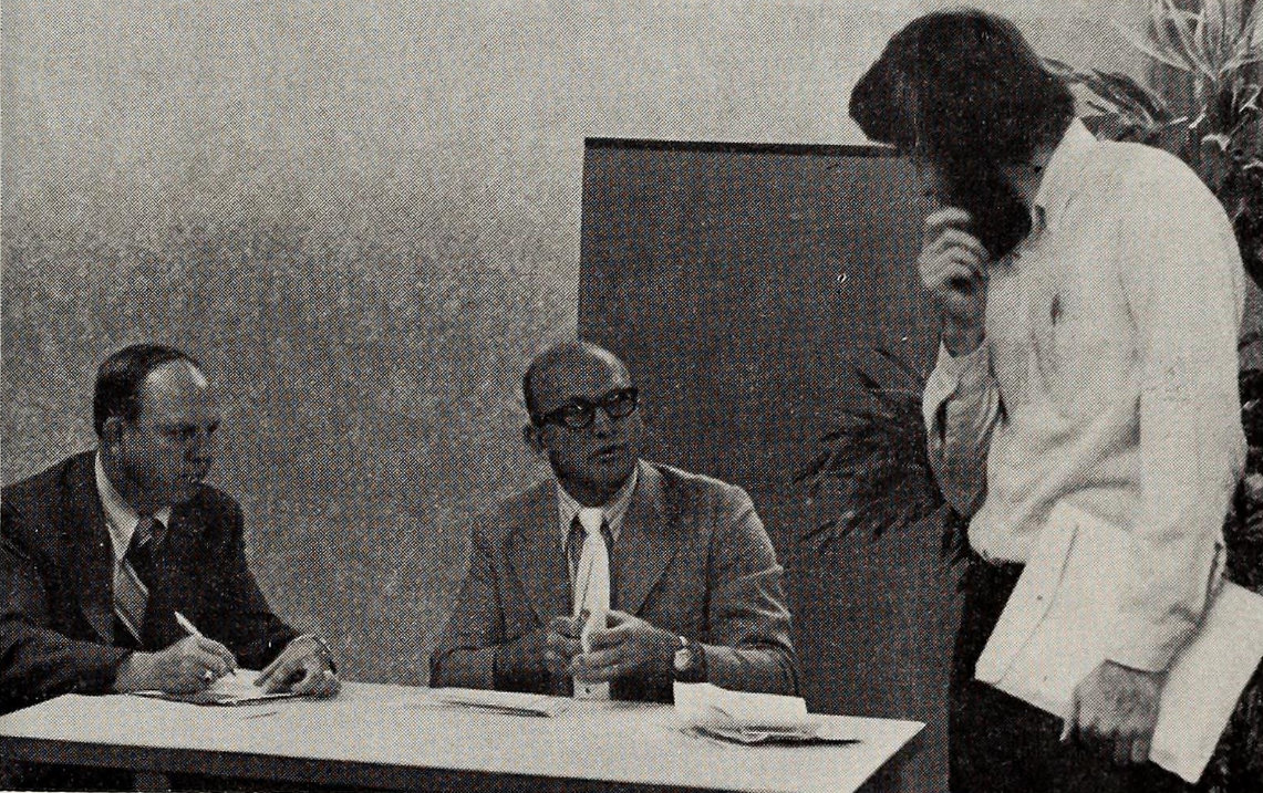 David Janzen, standing at right, talks with two Internal Revenue Service officials, seated behind a desk to the left