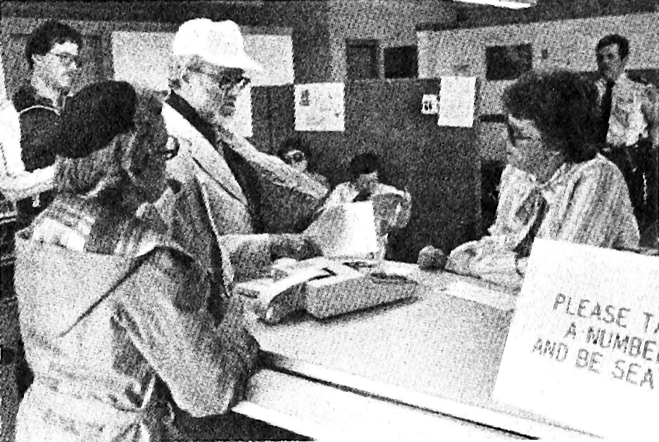 Two people are speaking with a third at an office counter. An adding machine is on the countertop along with a sign that reads “please take a number and be seated.”