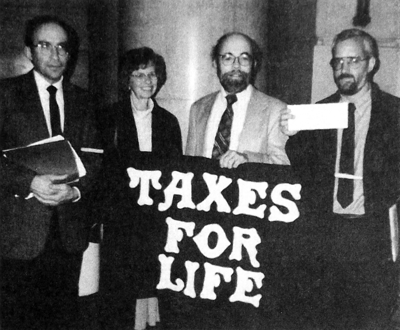 Herb Myers, Annn Marie Judson, John Stoner, and Dave Schrock-Shenk, facing the camera, stand behind a “Taxes For Life” banner. Schrock-Shenk holds up a check.