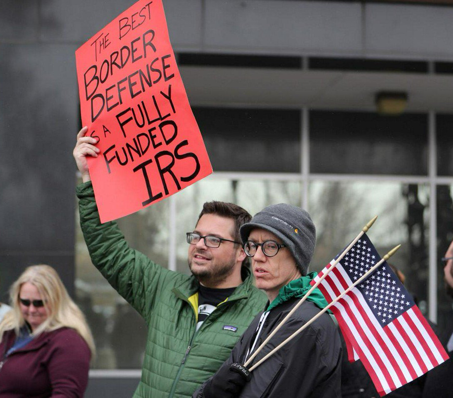 Three people stand in front of a building, one holds two American flags, another holds a sign that reads “The best border defense is a fully funded I.R.S.”