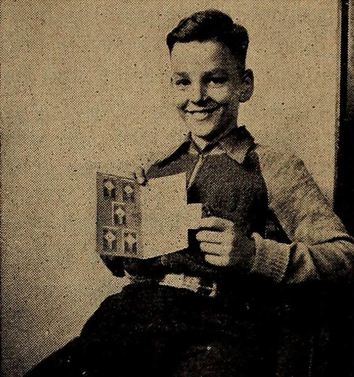 a seated boy smiles toward the camera as he holds his Brethren Service Stamps card with five stamps on it