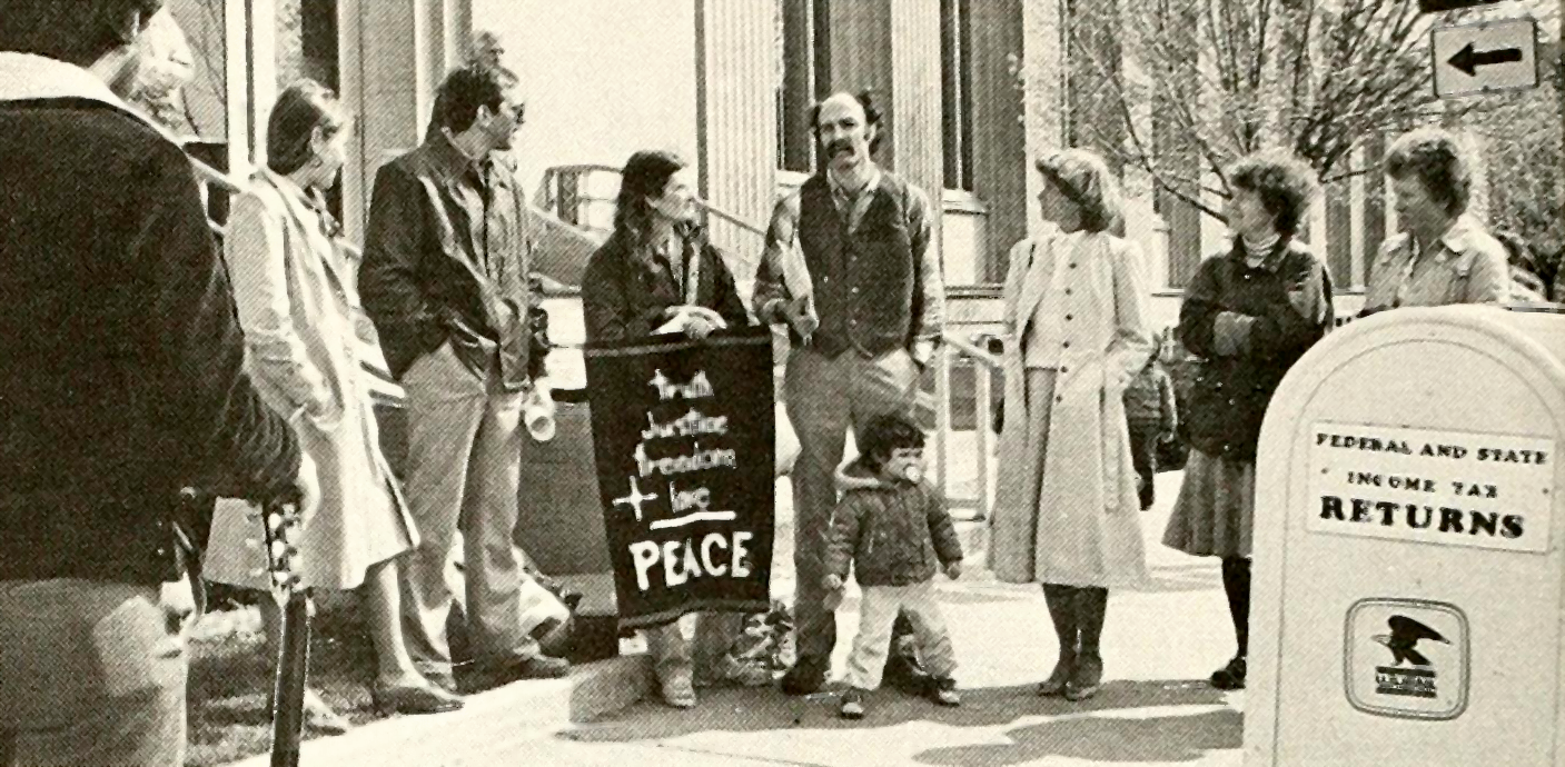 about ten people stand in a circle on the sidewalk, one holding a banner reading “truth + justice + freedom + love = peace” while in the foreground is a mailbox labeled “Federal and State Income Tax Returns”