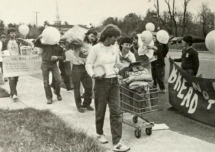 Several people walking along a sidewalk, some with banners, some with balloons, one hoisting a large bag on his shoulders, one pushing a shopping cart, some holding banners
