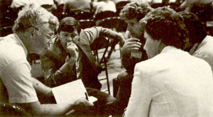 five people sit in a circle looking at each other, one holding a book, with folding chairs in the background