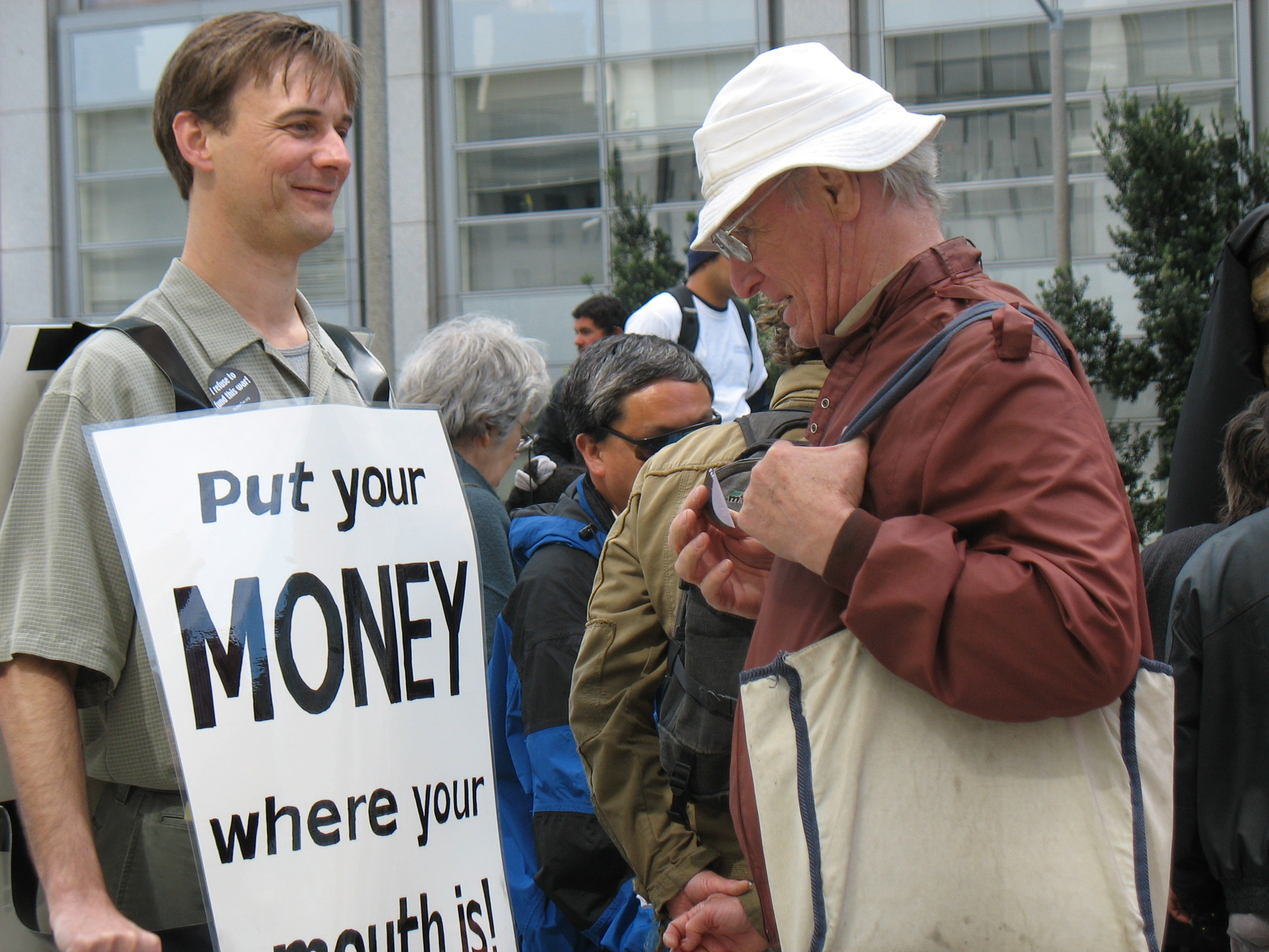 A rally attendee reads my “Put Your Money Where Your Mouth Is” sandwich board at an earlier rally.